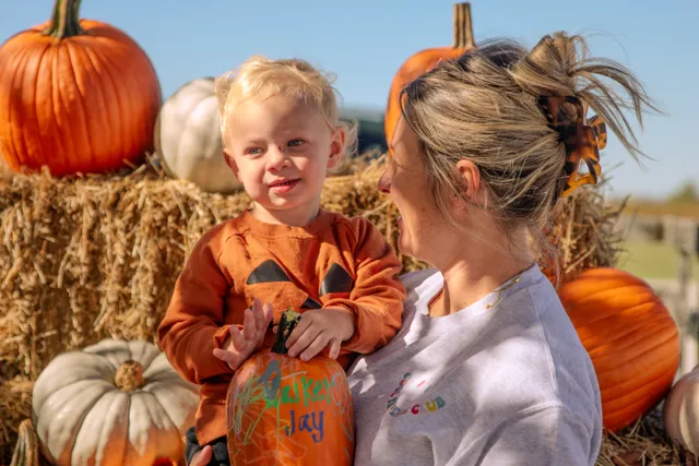 Blond toddler in an orange jack-o'-lantern sweatshirt holds a decorated pumpkin beside a smiling woman near hay bales