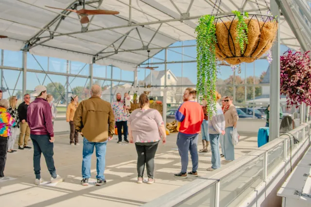 Group of people standing in a circle inside a sunny glass greenhouse with hanging plant baskets and ceiling fans