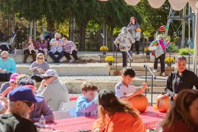 Kids decorate pumpkins at a red-checkered table while people sit on sunny stone steps with yellow mums behind them.