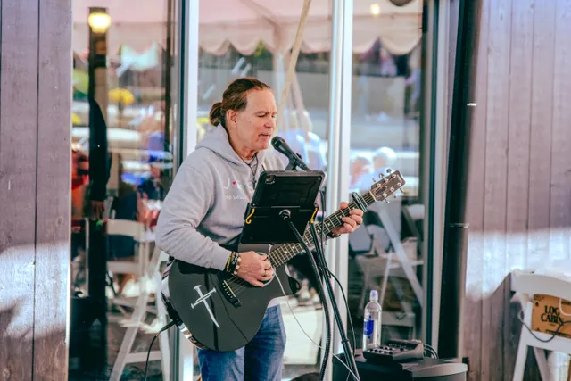Musician in a gray hoodie sings into a microphone while playing a black acoustic guitar with a white sword design