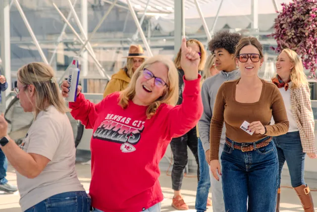 Smiling woman in a red Kansas City Chiefs shirt raises a fist and a can while dancing with others in a greenhouse