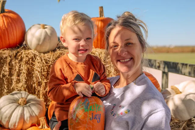 Smiling woman and toddler in orange sweater holding a decorated pumpkin beside hay bales and pumpkins