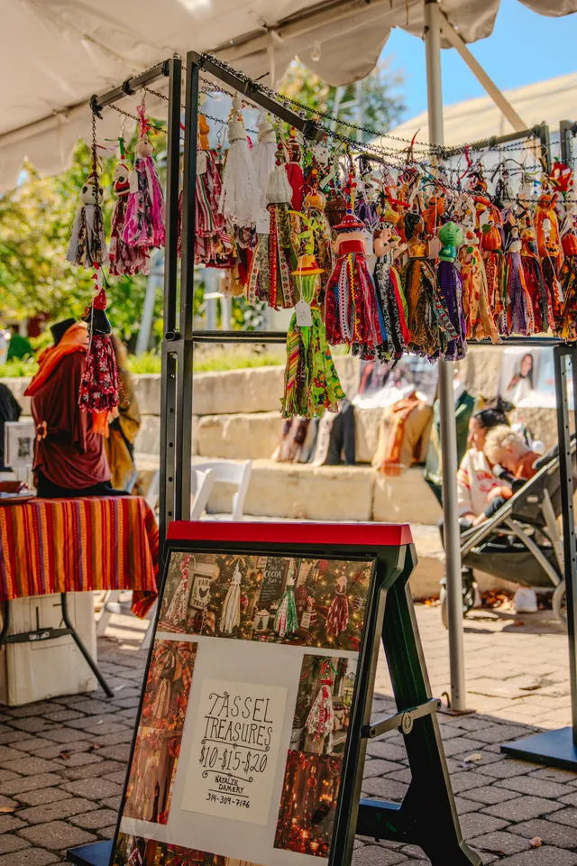 Rack of colorful fabric tassel dolls hanging under a white tent, with a framed Tassel Treasures price sign below