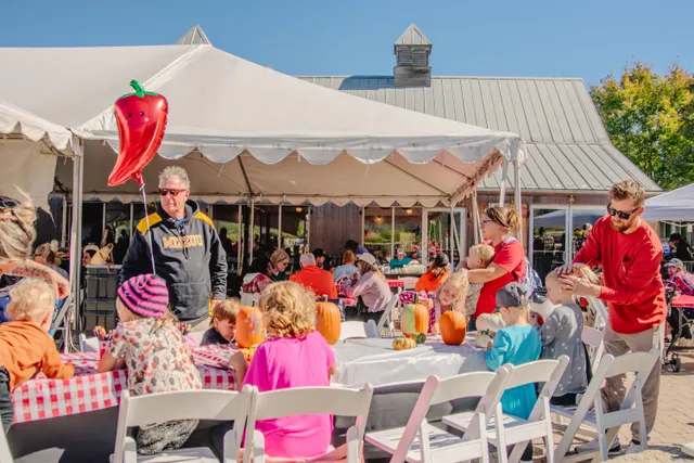 Children paint pumpkins at outdoor tables under a white tent as adults watch, with a red chili pepper balloon overhead.