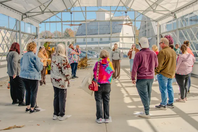 Visitors stand in a circle inside a sunny open greenhouse with ceiling fans, cornstalks, pumpkins and a white barn behind