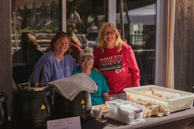 Three people smile behind a table with black soup warmers, a tip cup and small cups of chili toppings at an outdoor booth.