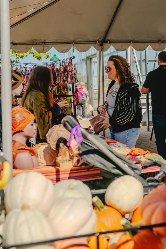 Shopper in sunglasses holding a pink wallet talks with a vendor under a white tent, white and orange pumpkins in front