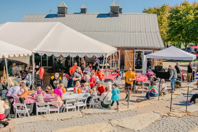 Families at tables under white tents decorating pumpkins beside a gray metal-roofed barn on a sunny day