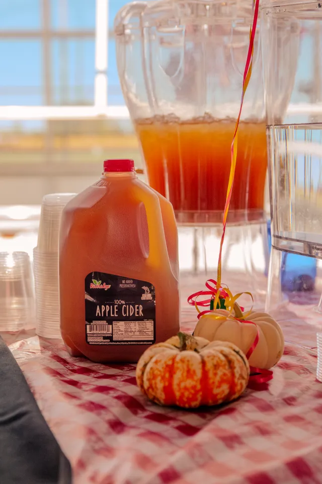 Gallon jug of apple cider beside mini pumpkins and a drink dispenser on a red gingham tablecloth