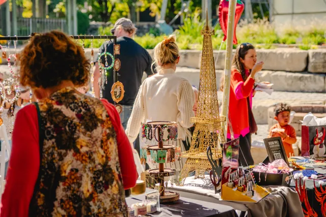 Shoppers browse outdoor vendor tables with hanging jewelry, beaded bracelets and a gold Eiffel Tower model on a sunny day.