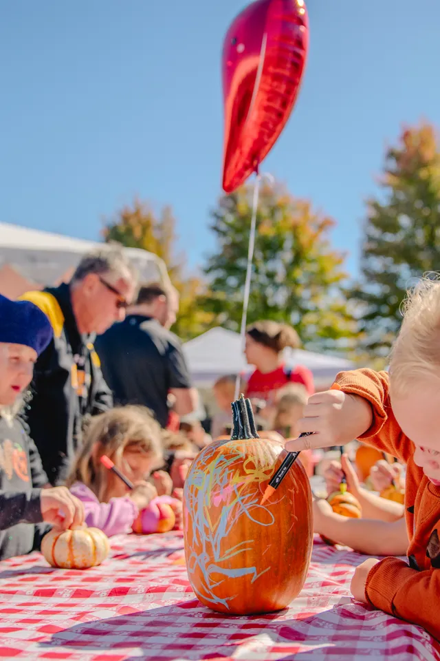 Child in an orange sweater colors a pumpkin with markers at a red gingham table as other kids decorate pumpkins