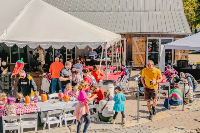 Families gather at red-checkered tables under white tents beside a wooden barn, with kids decorating small pumpkins.