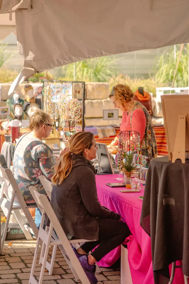 Two women seated in white folding chairs at a pink-draped vendor table with beaded jewelry displays under a white tent.