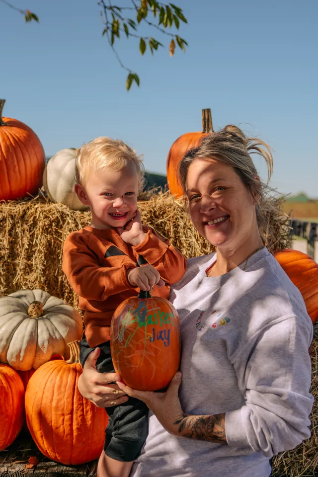 Smiling woman holds a blond toddler in an orange jack-o'-lantern sweatshirt and a decorated pumpkin before hay bales.