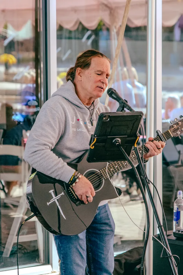 Man in gray hoodie sings into a microphone while playing a black acoustic guitar with a white nail cross design