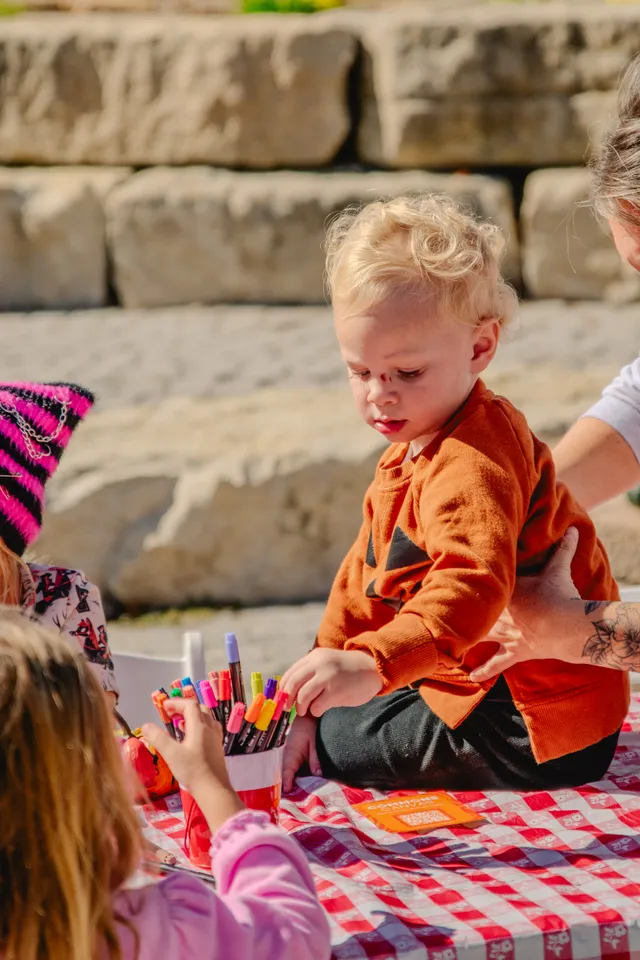 Curly-haired toddler in an orange jack-o'-lantern sweater sits on a red checkered table reaching for a cup of colorful markers.