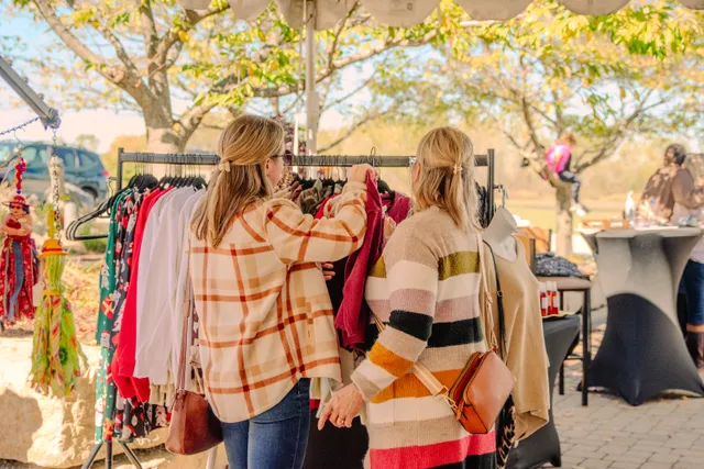 Two women seen from behind browse a clothing rack at an outdoor vendor booth under autumn trees.