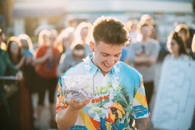 Smiling young man in a bright Hawaiian-style shirt and white lei holds a clear box of cash before a crowd at sunset