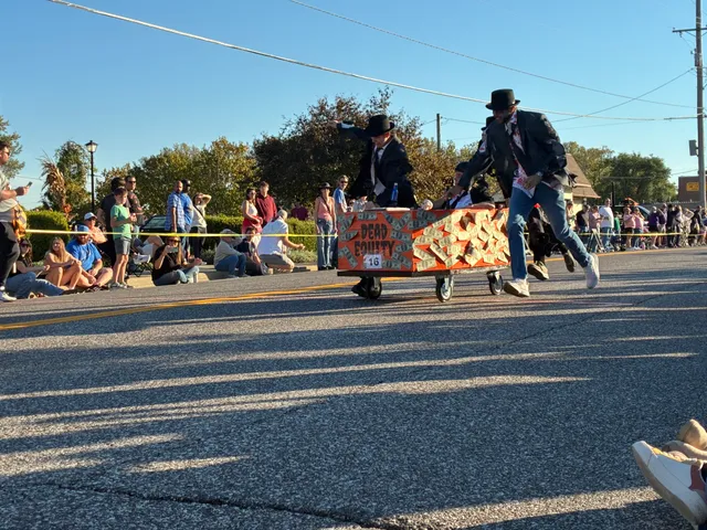 Racers in black top hats and suits push an orange coffin on wheels, covered in fake money and labeled Dead Equity, No. 16.