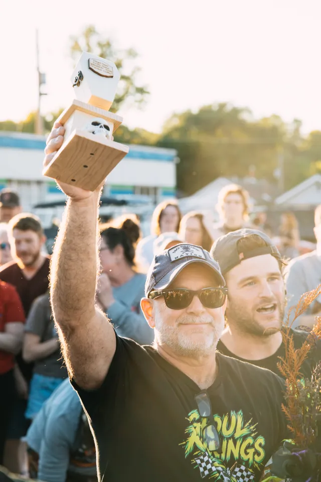 Man in cap and sunglasses raises a wooden coffin-and-skull trophy above a cheering crowd outdoors on a sunny day