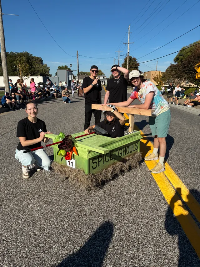Five people pose with a green coffin cart labeled Spice Ghouls, number 11, decorated with vegetables, on a street.