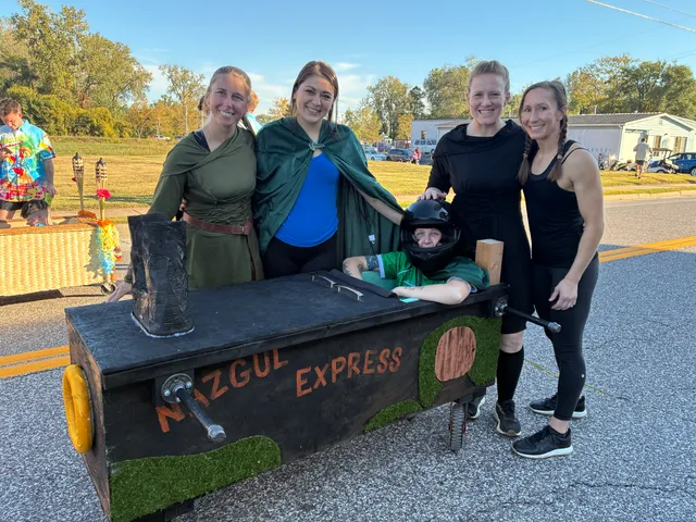 Four women in green cloaks and black outfits pose around a black wheeled coffin labeled Nazgul Express with a helmeted driver inside.