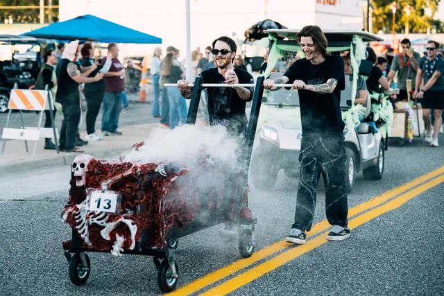 Two men in black push a smoking coffin cart decorated with skeletons and red gore, number 13, down a street
