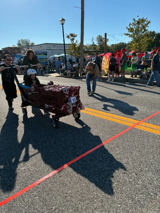 Two people push a skeleton-decorated red coffin cart number 13 with a helmeted rider toward a red finish line on a street.