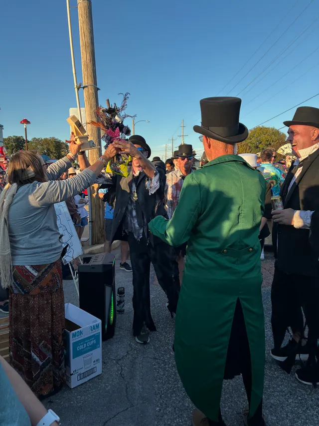 Woman hands a trophy and spooky bouquet to a man in a tattered black suit as men in top hats and a green tailcoat watch