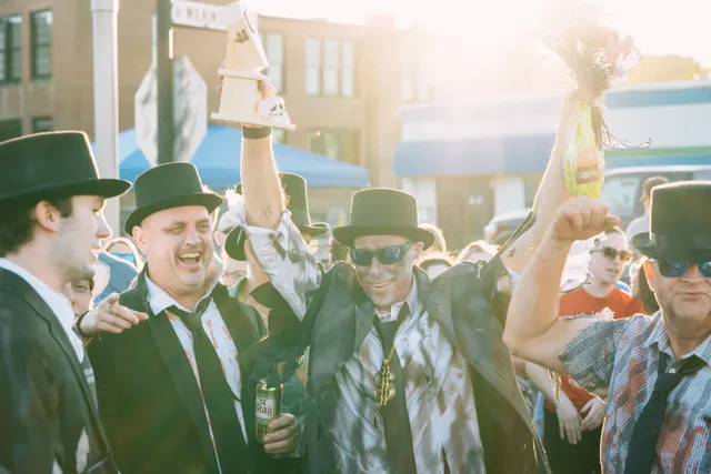 Men in black top hats and fake-bloodied shirts cheer as one raises a trophy and another lifts a bouquet on a city street