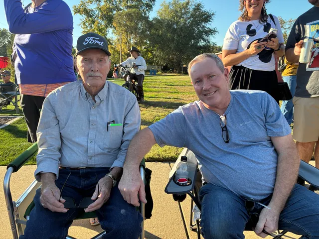 Older man in a Ford cap and younger man in a gray T-shirt sit in folding camp chairs on a sunny lawn, smiling.