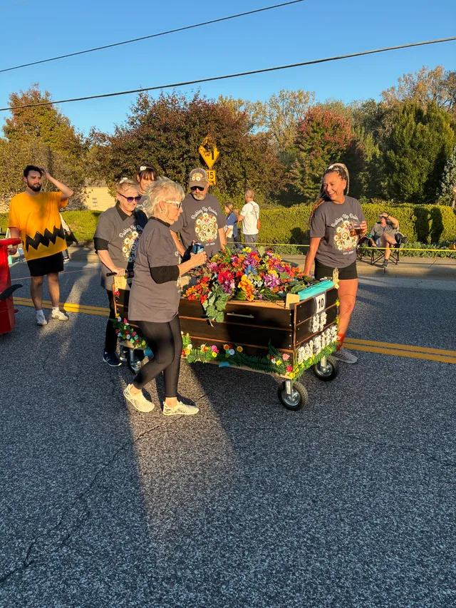 Team in matching gray sugar-skull shirts pushes a wheeled wooden coffin topped with bright flowers down a street