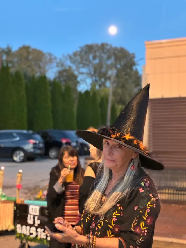 Woman in a tall black witch hat and floral black top under a full moon, another woman sipping a drink behind her