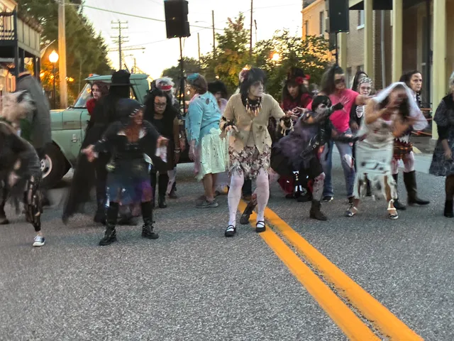 Crowd of people in zombie costumes and pale makeup dancing in a street with a double yellow line, vintage green truck behind.