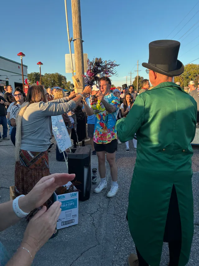 Man in a bright tropical shirt holds up a trophy topped with dark flowers as a crowd claps; man in green tailcoat and top hat watches