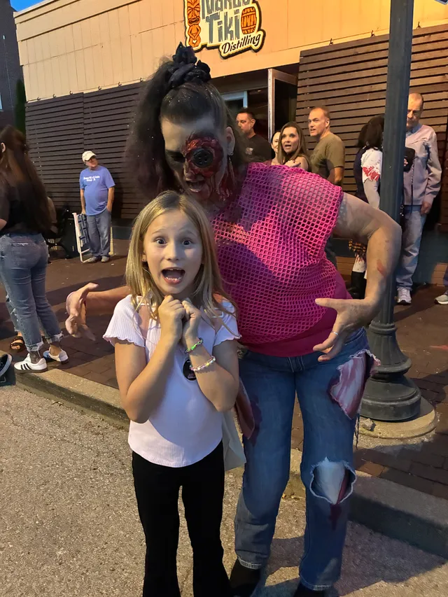 Girl in white top makes a mock-scared face as a zombie in pink mesh top and torn bloody jeans looms behind her on a sidewalk.