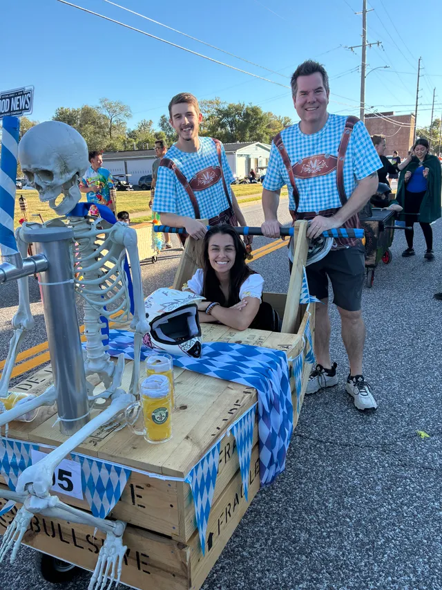 Two men in blue checkered lederhosen shirts push a wooden crate coffin with a smiling woman inside and a skeleton up front.