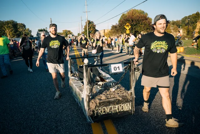 Two men in black Ghoul Runnings shirts push a coffin labeled Frenchtown with number 01 down a street lined with spectators.