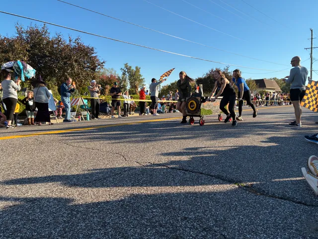Four women push a wheeled coffin cart down a street as spectators line the curb and a man waves a checkered flag.