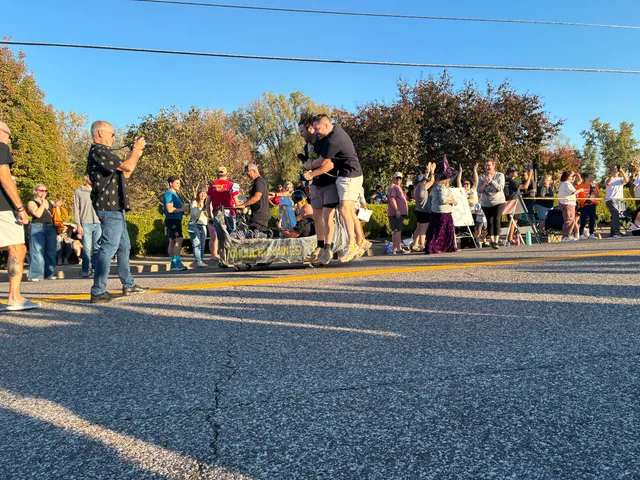 Two men in black shirts embrace and jump at a coffin race finish line as a crowd cheers and a man takes photos