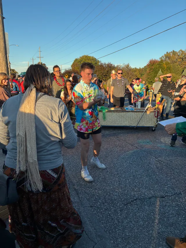 Man in a bright tropical shirt and lei holds a glass trophy on a street, with a tiki-themed coffin cart and crowd behind