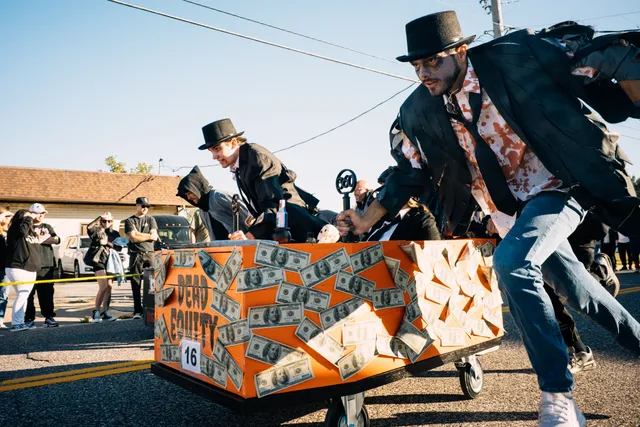 Runners in top hats and fake-blood shirts push an orange coffin covered in fake $100 bills, numbered 16, down a street.