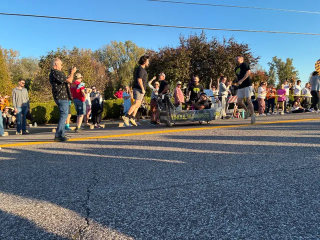 Team in black shirts pushes a wheeled coffin with a rider past cheering spectators on a sunny street.