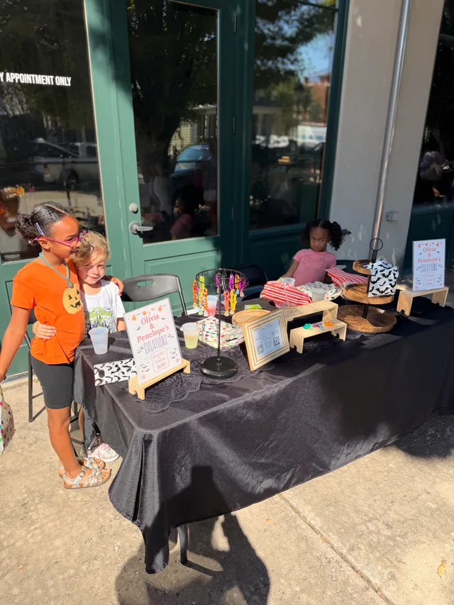 Two kids hug beside a black-draped table with beaded bracelets and Halloween cookie bags; another child sits behind it.