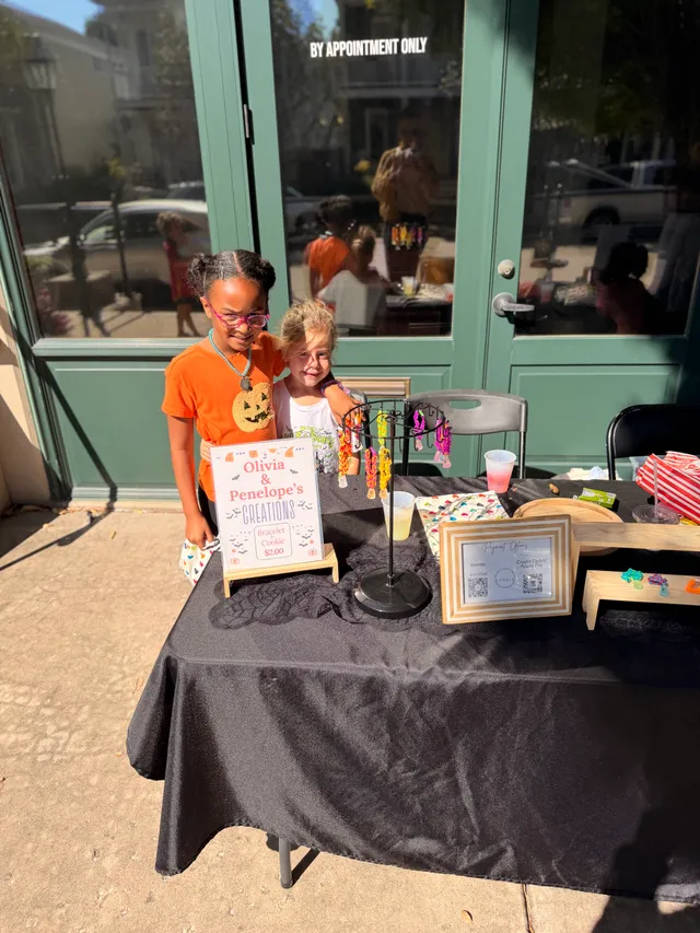 Two girls stand behind a black-covered table with a sign reading Olivia & Penelope's Creations and a bracelet display.