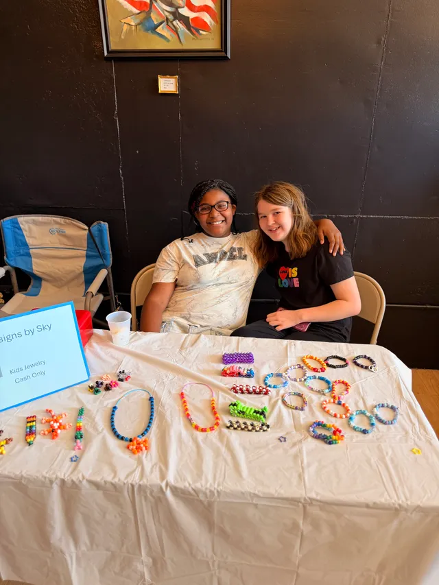 Two girls smile behind a white-covered table displaying colorful beaded bracelets, necklaces and keychains for sale.
