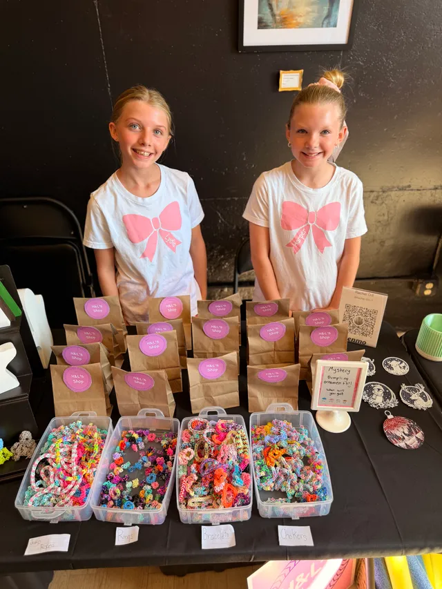 Two girls in matching white shirts with pink bows stand behind a table of rubber band bracelets and brown paper bags.