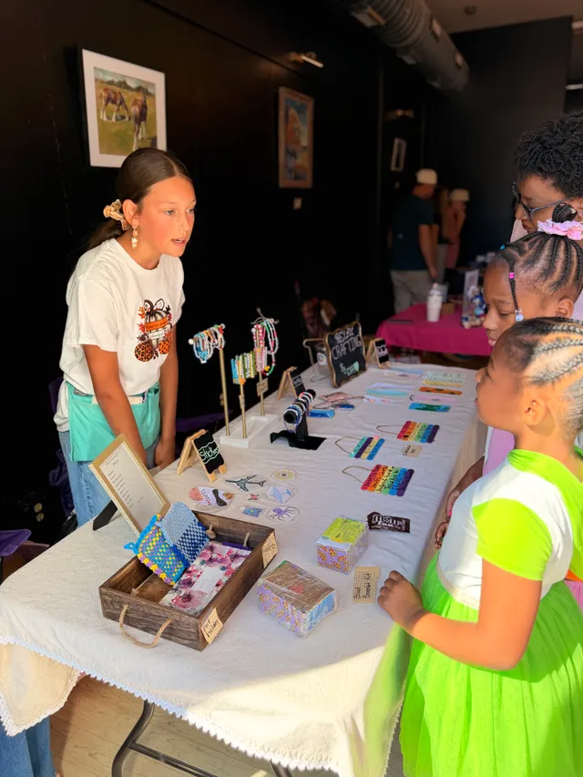 Girl in a pumpkin T-shirt and teal apron talks with young shoppers at a table of handmade bracelets, stickers and crafts.