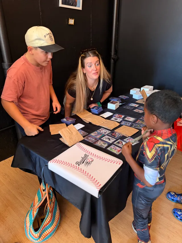 Boy in a cap and woman sell baseball cards at a table as a child in a red-and-gold superhero suit holds cash