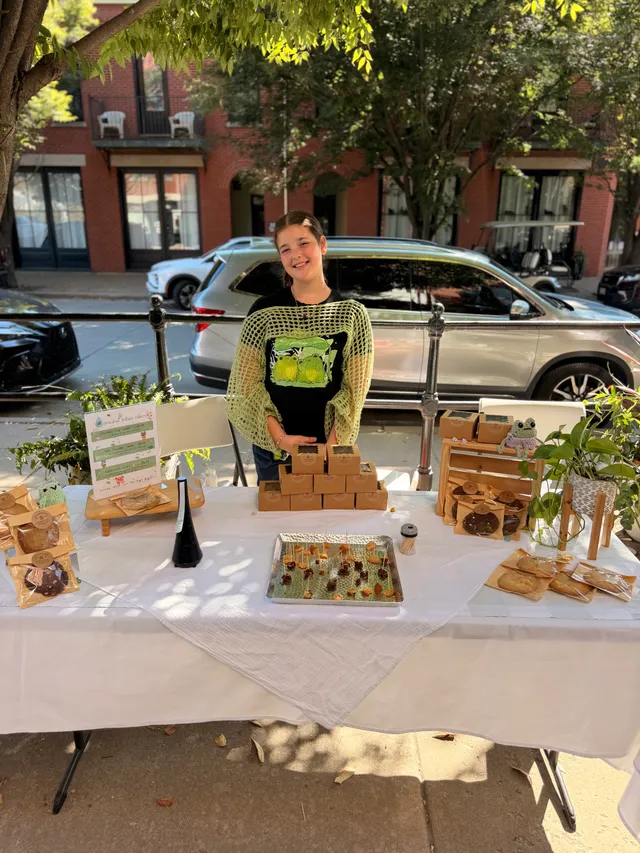 Young vendor in a green crochet top and frog T-shirt smiles behind a table of bagged cookies, kraft boxes and samples.